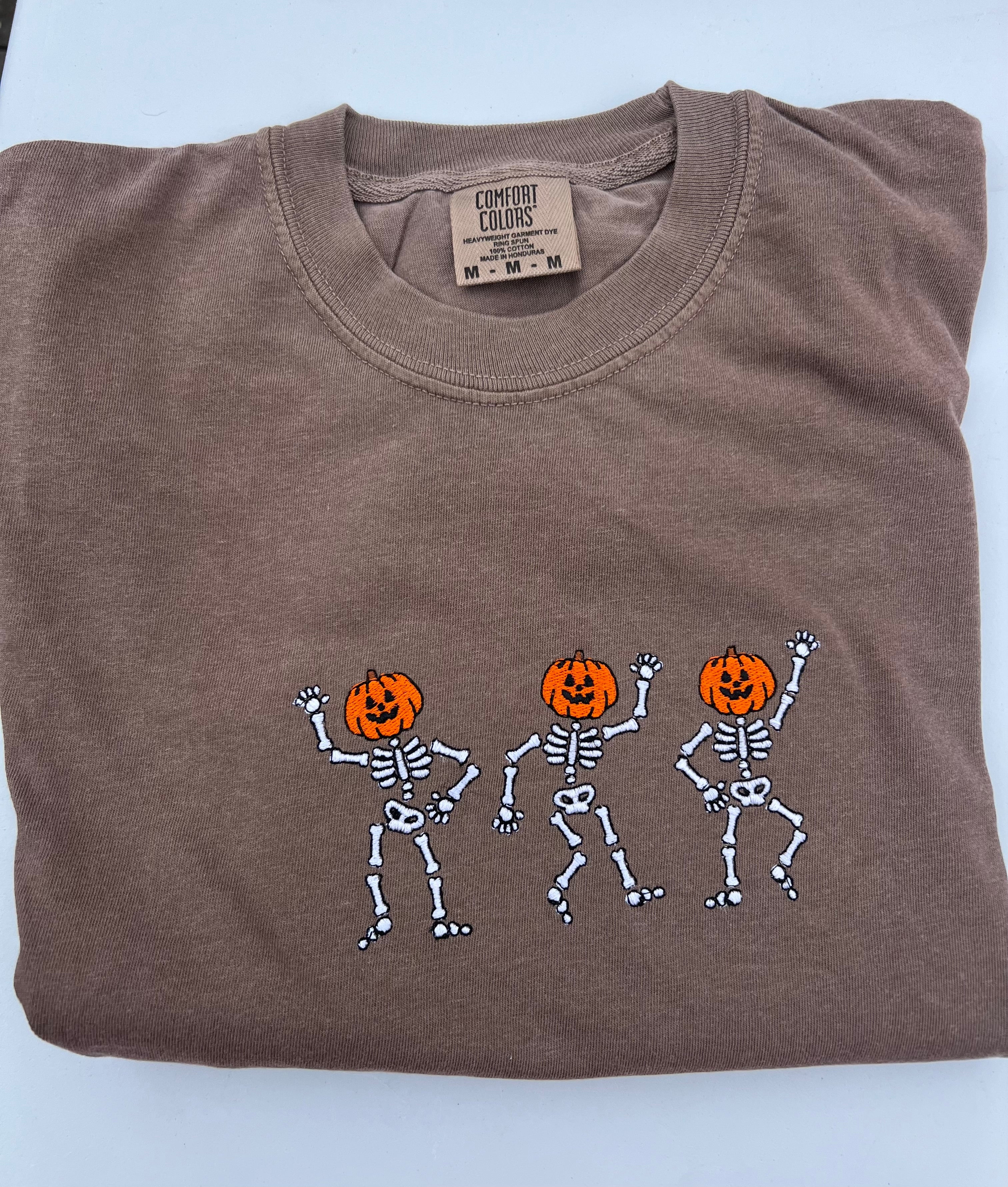 A folded brown comfort color tshirt embroidered with the image of three dancing skeleton pumpkins on the front