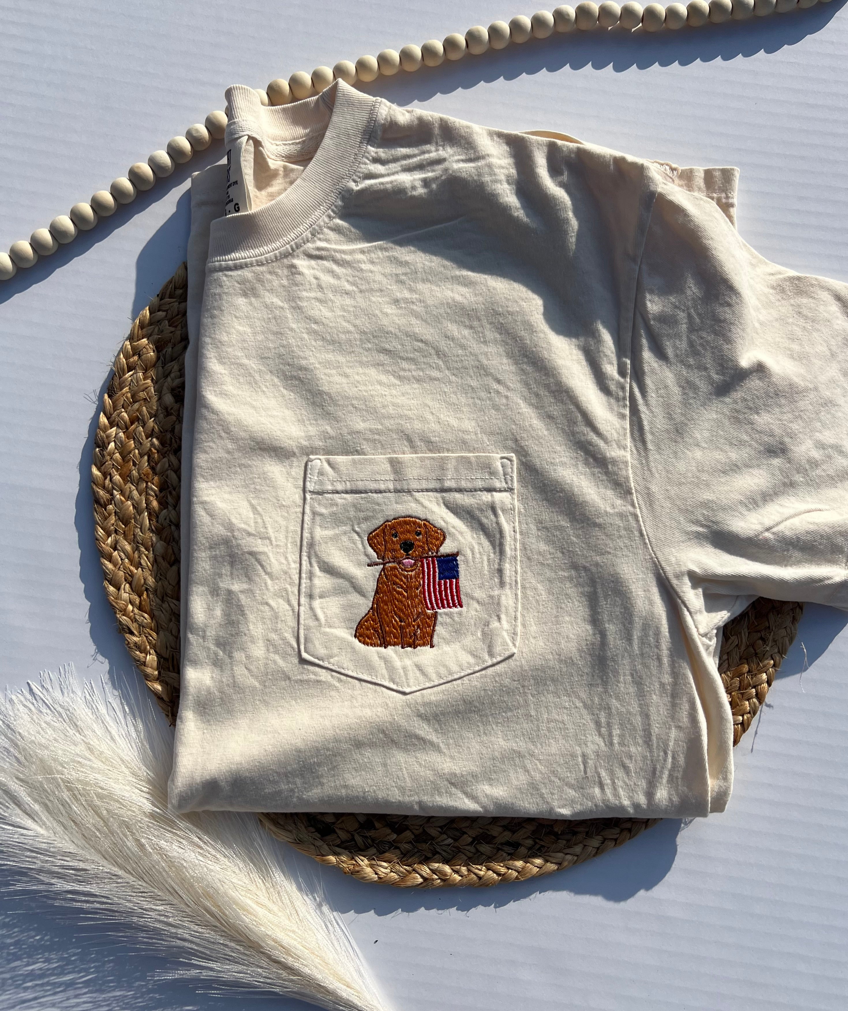Folded beige t-shirt with a pocket featuring an embroidered dog and American flag design on a textured surface.