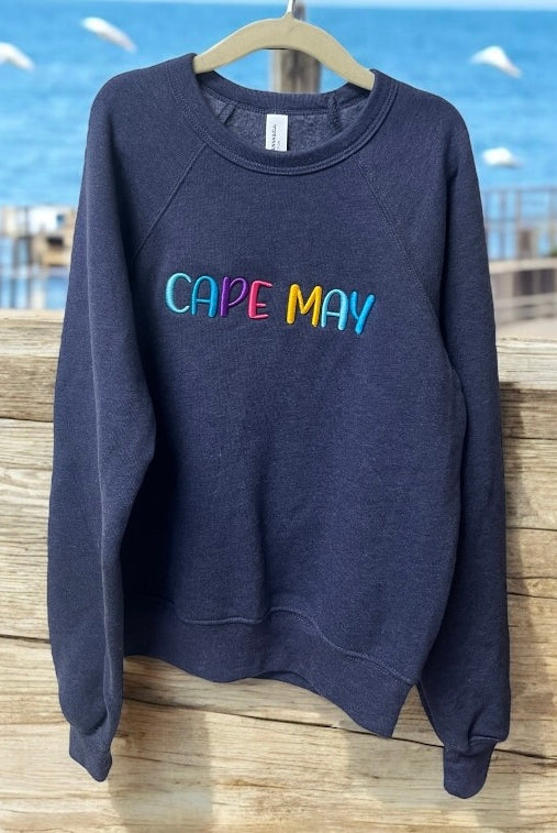 Navy blue sweatshirt with 'CAPE MAY' in colorful letters on a wooden surface with a beach background.