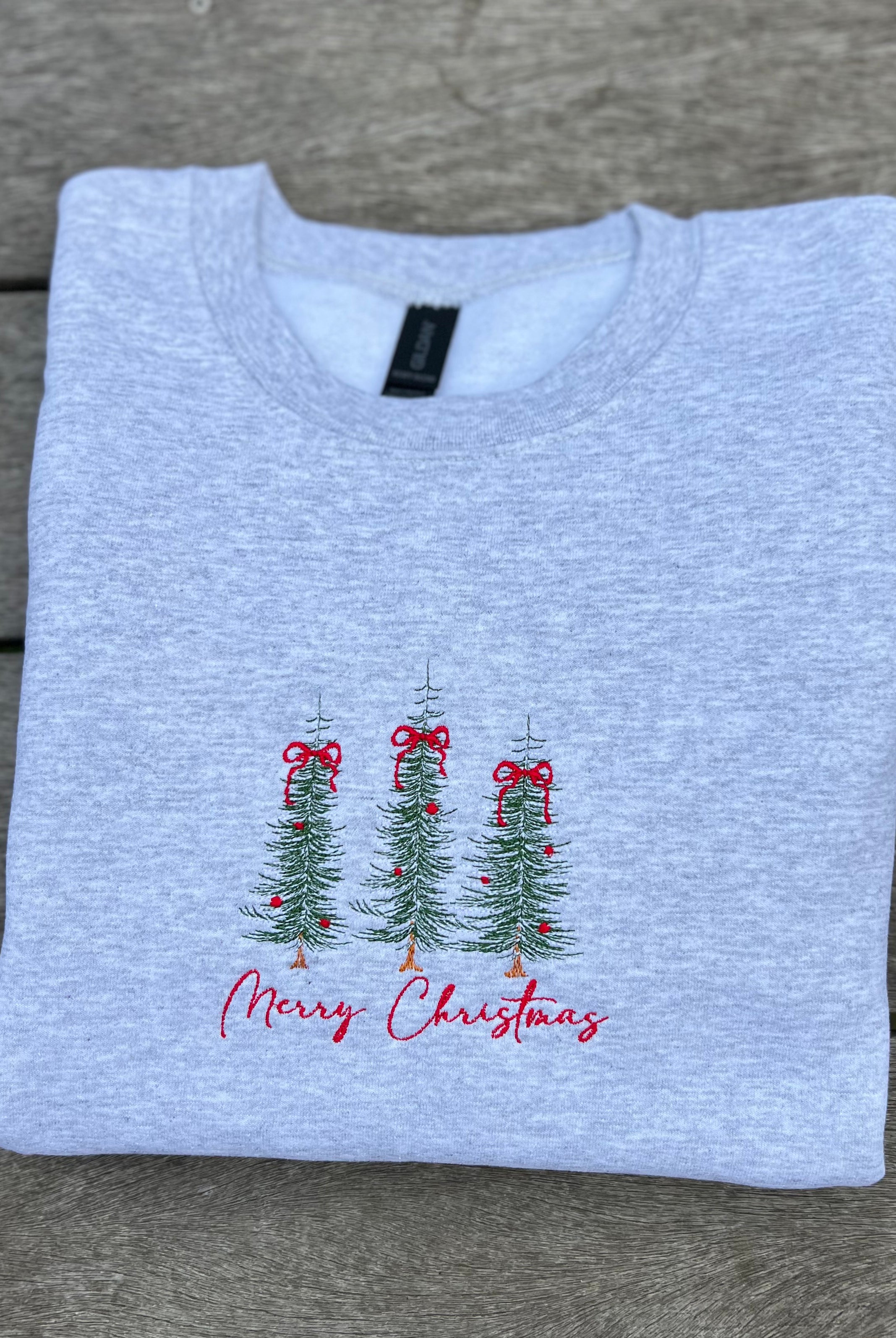 Folded light gray sweatshirt with Christmas tree and bow that says merry christmas on a wooden surface