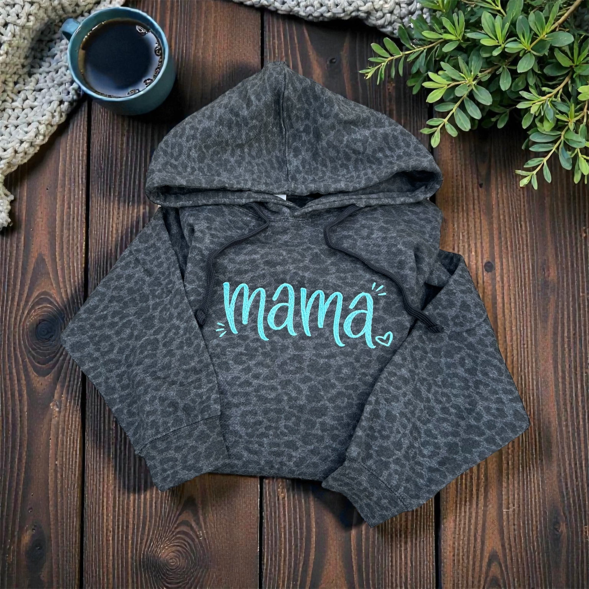 Gray leopard print hoodie with 'mama' text on a wooden surface with a cup and plant.
