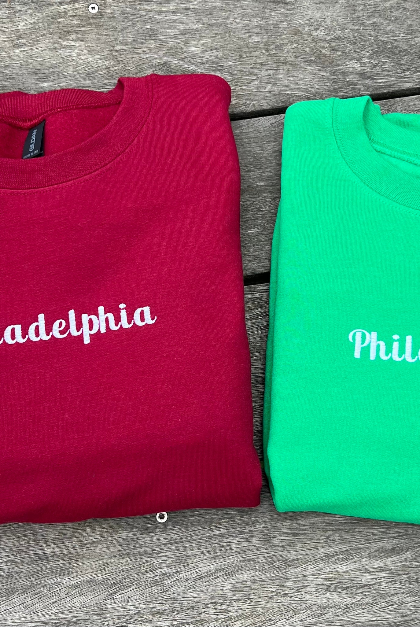 Two folded sweatshirts, one red and one green, with 'Philadelphia' embroidred on them