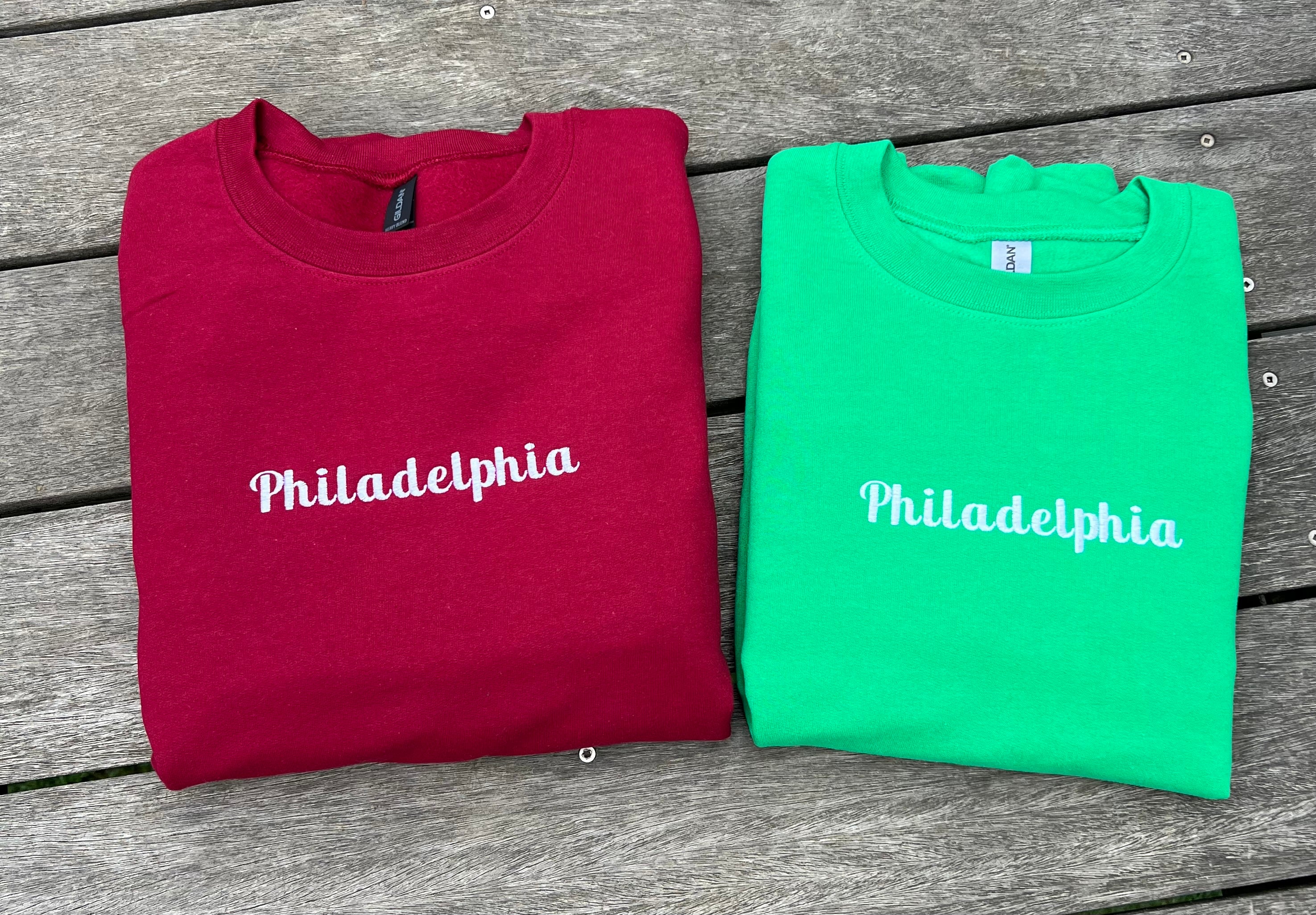 Two folded sweatshirts, one red and one green, with 'Philadelphia' embroidred on them