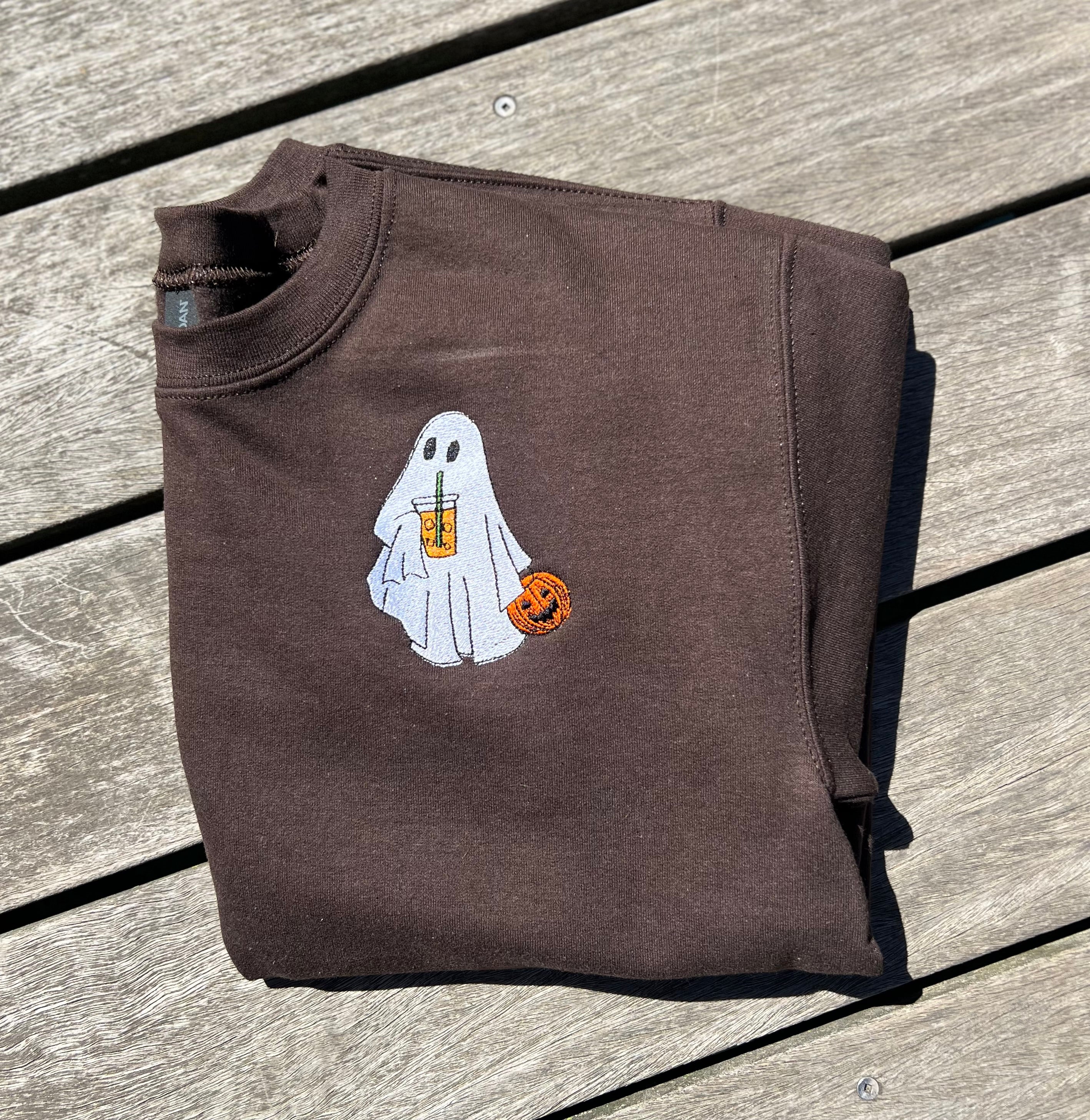 Folded brown sweatshirt with a ghost holding an iced coffee and halloween basket, embroidered