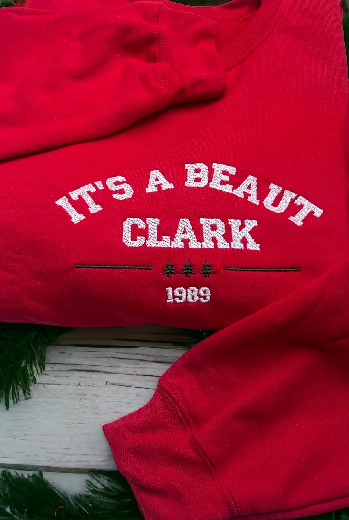 Red sweatshirt with 'It's a Beaut Clark 1989' text on a wooden surface with greenery.