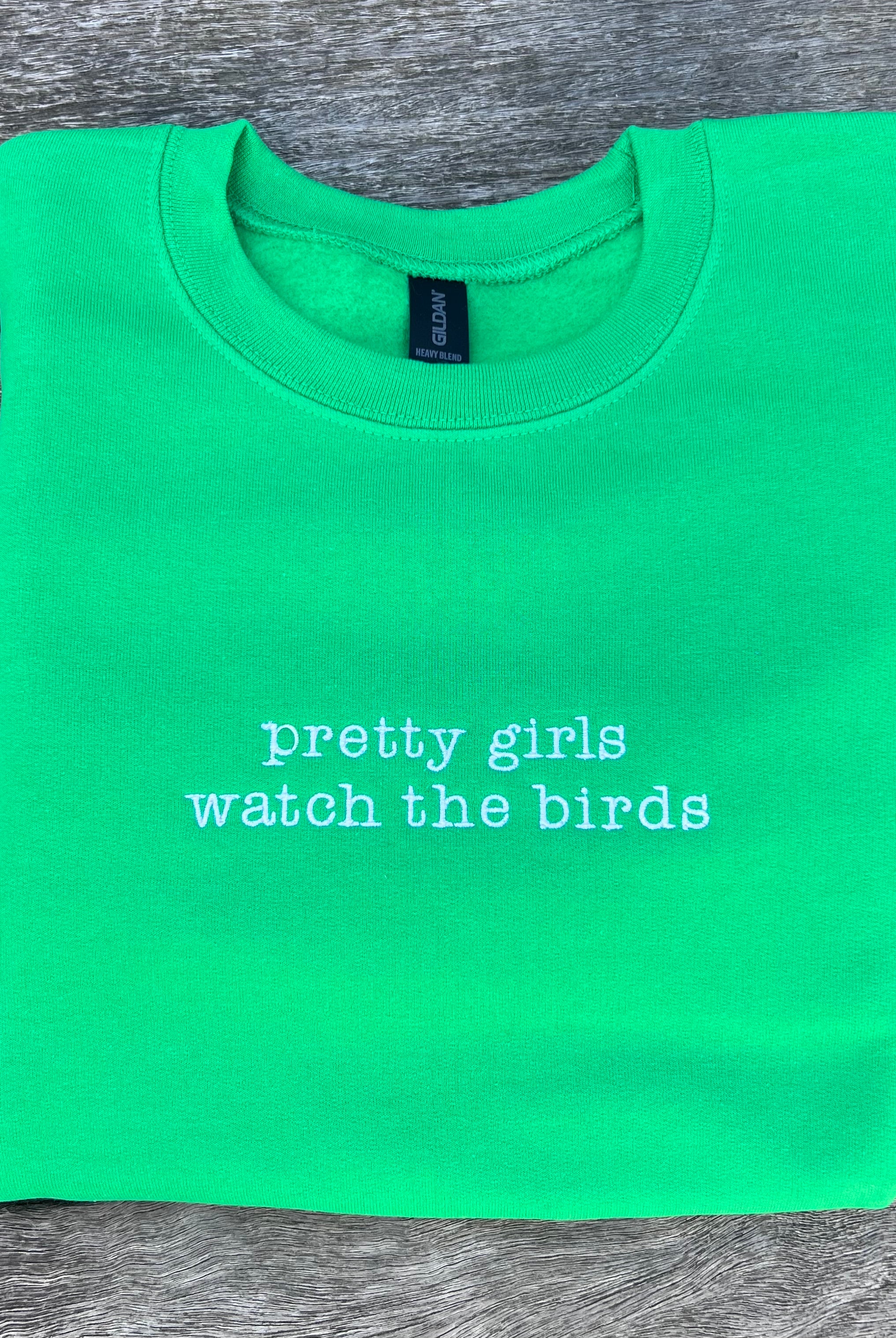 Green sweatshirt with 'pretty girls watch the birds' text on a wooden surface