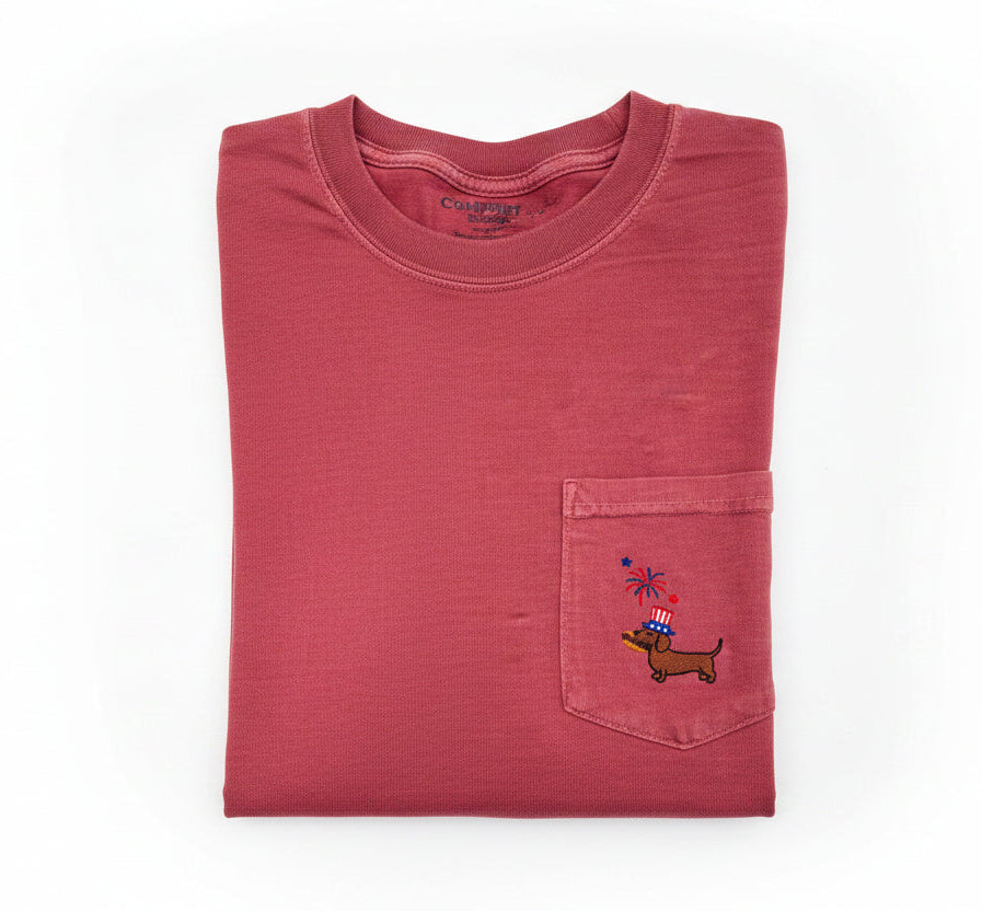 Folded red shirt with a patriotic dachsund eating a hotdog on a pocket design on a white surface