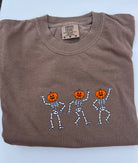 A folded brown comfort color tshirt embroidered with the image of three dancing skeleton pumpkins on the front
