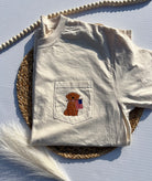 Folded beige t-shirt with a pocket featuring an embroidered dog and American flag design on a textured surface.