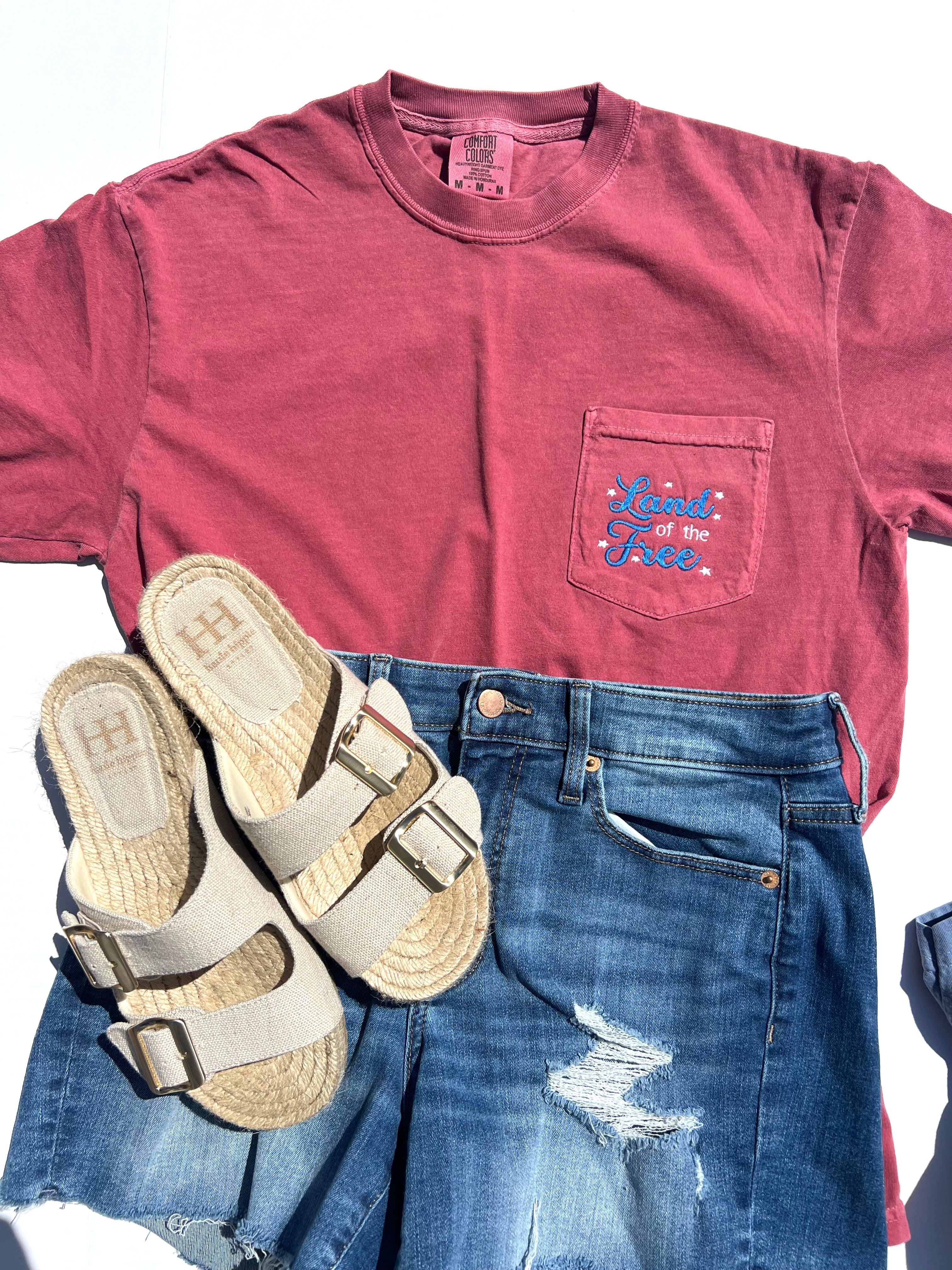 Red t-shirt with land of the free embroidered on a pocket design, blue jeans, and sandals on a white background