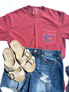 Red t-shirt with land of the free embroidered on a pocket design, blue jeans, and sandals on a white background