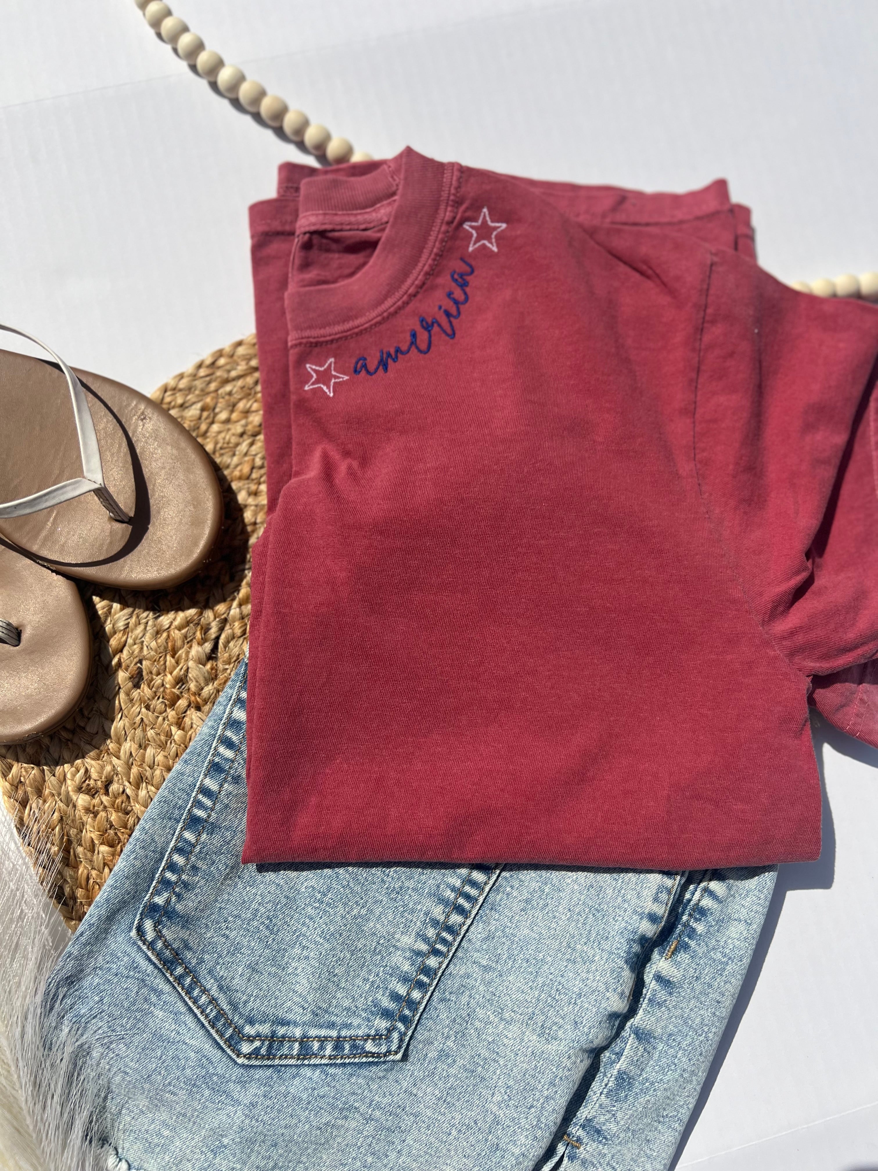 Red sweatshirt with 'America' text and star design on a white surface with denim jeans and sandals.
