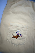 Beige shirt with a pocket featuring an embroidered dachshund and flag design on a yellow shirt