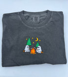 A folded dark gray crewneck t-shirt featuring a cute cartoon graphic of two white ghosts roasting marshmallows by a campfire under green pine trees and a crescent moon.