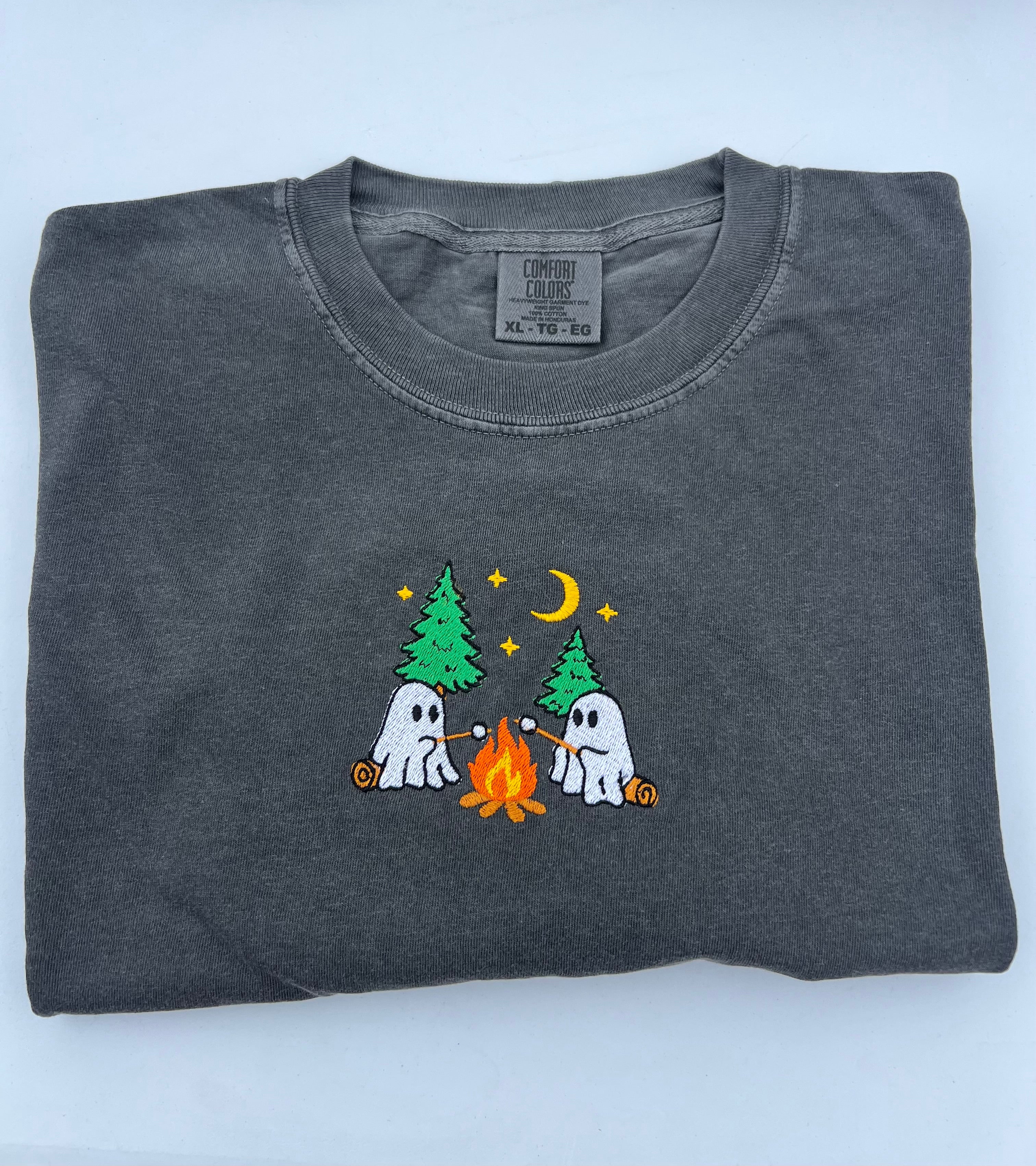 A folded dark gray crewneck t-shirt featuring a cute cartoon graphic of two white ghosts roasting marshmallows by a campfire under green pine trees and a crescent moon.