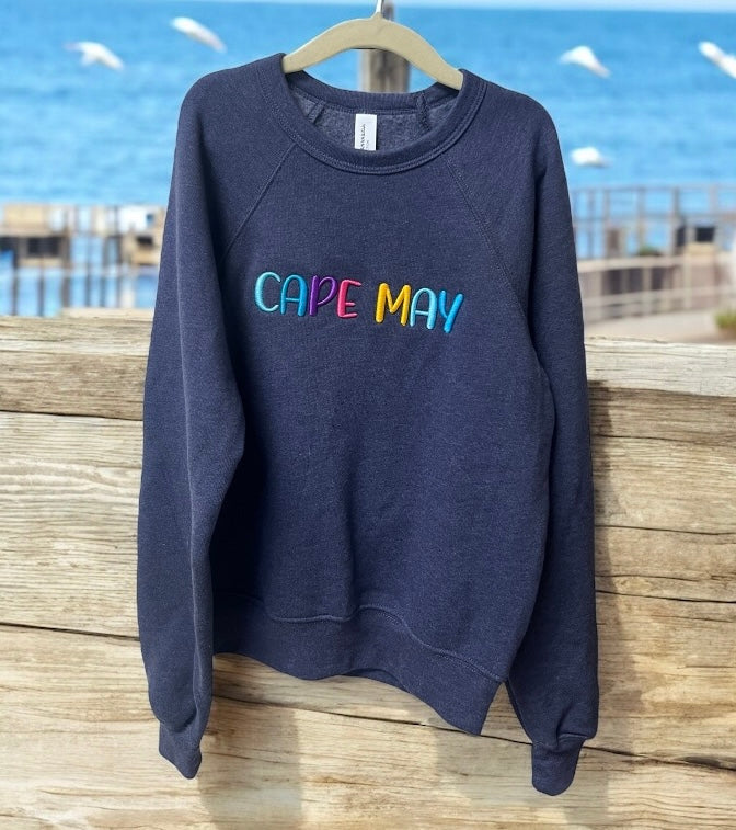 Navy blue sweatshirt with 'CAPE MAY' in colorful letters on a wooden surface with a beach background.