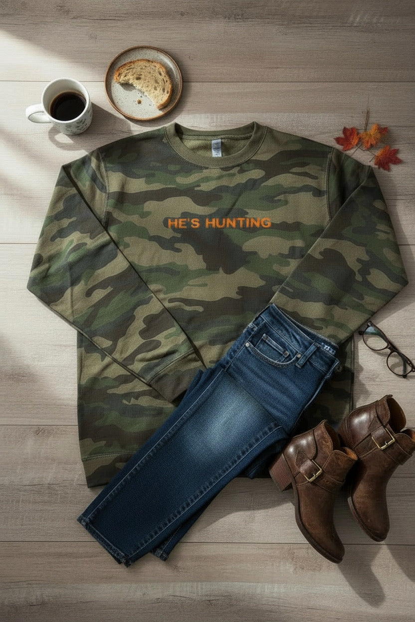 Camouflage sweatshirt with 'HE'S HUNTING' text, blue jeans, brown boots, and a coffee cup on a wooden surface.