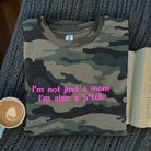 Camouflage t-shirt with pink text on a textured surface with a cup of coffee and book.