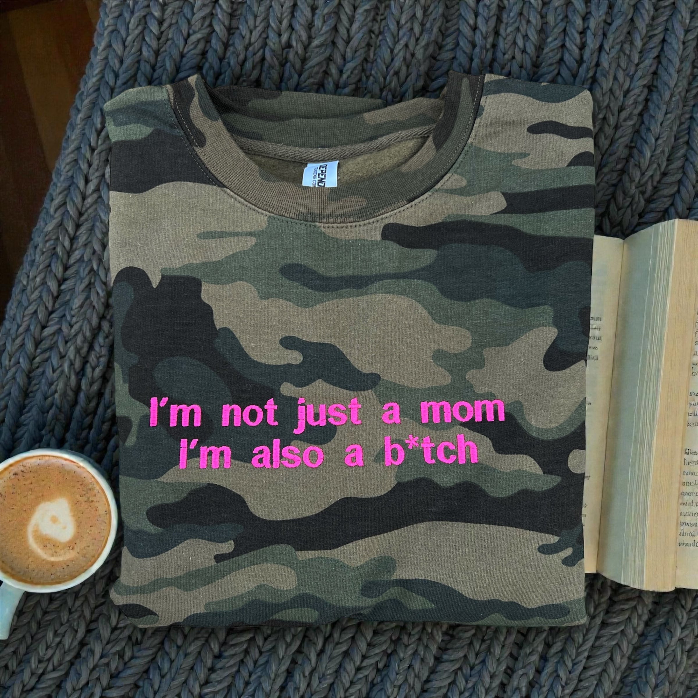 Camouflage t-shirt with pink text on a textured surface with a cup of coffee and book.