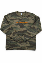 Camouflage long-sleeve shirt with 'HE'S HUNTING' text on a white background