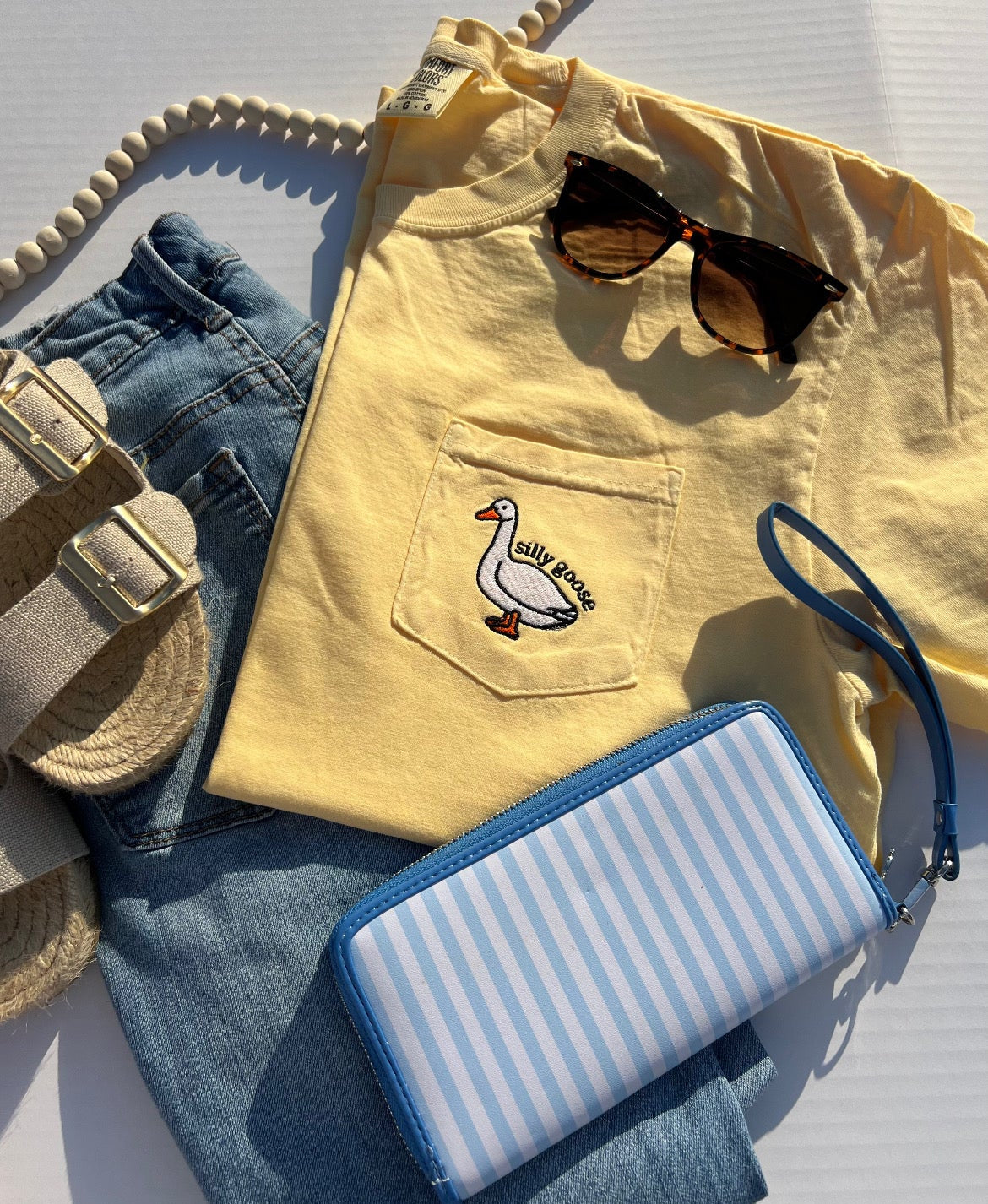 Yellow shirt with a duck design, sunglasses, blue jeans, and a striped wallet on a light gray surface.