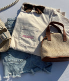 Light-colored shirt with 'mama' embroidery, denim shorts, straw bag, and sunglasses on a white surface.