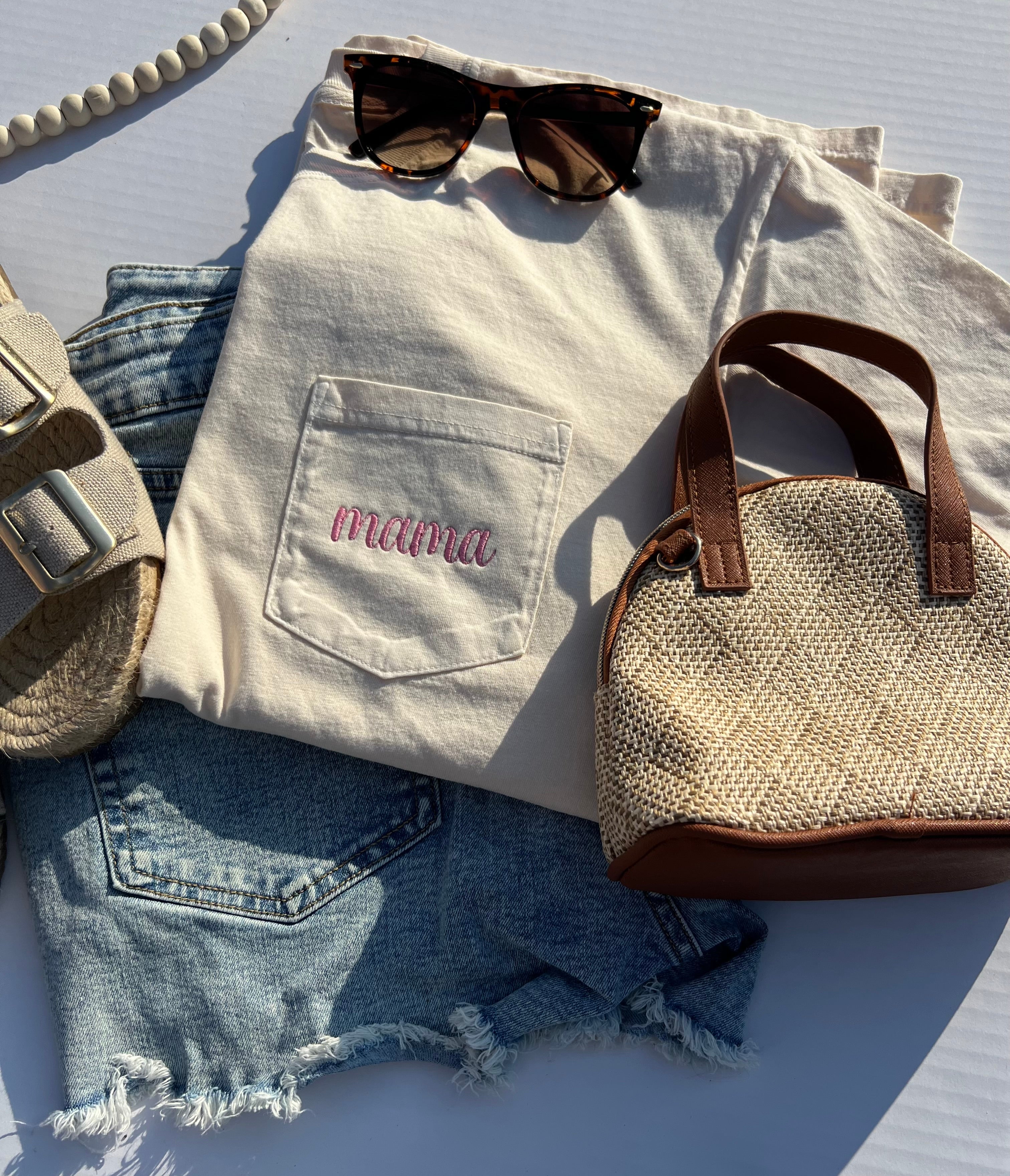 Light-colored shirt with 'mama' embroidery, denim shorts, straw bag, and sunglasses on a white surface.