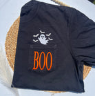 A black short-sleeve T-shirt laid flat on a woven placemat, featuring a chest pocket with orange embroidered 'BOO' lettering and a white ghost with bats above it.
