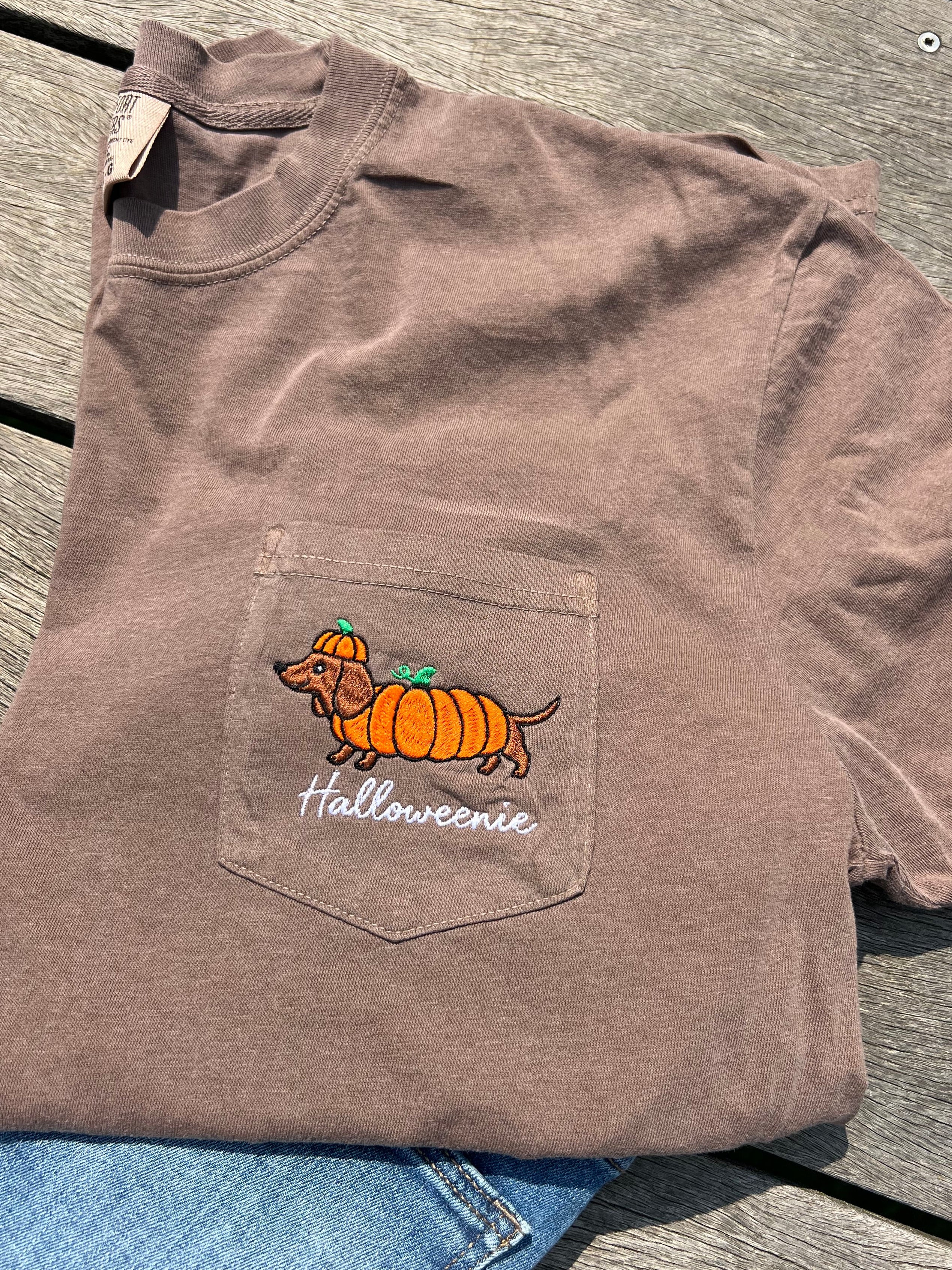 A brown pocket t-shirt with a graphic of a dachshund wearing an orange pumpkin hat and the text 'Halloweenie' on the front pocket.