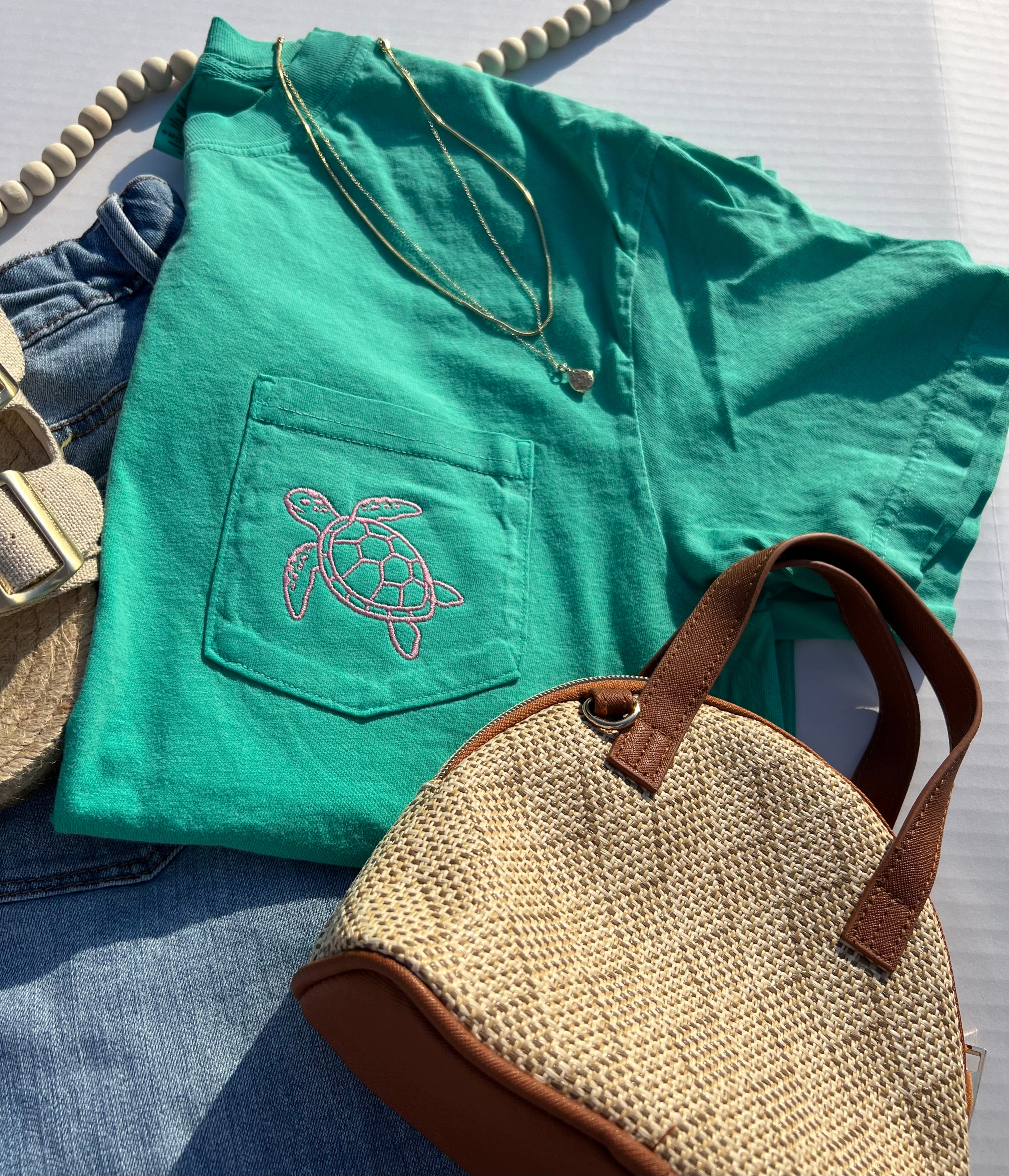 Green t-shirt with turtle design, straw bag, and sandals on a light surface