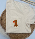 An off-white short-sleeve t-shirt with a front pocket featuring an embroidered brown puppy holding an orange pumpkin, styled on a woven placemat with wooden beads.
