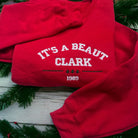 Red sweatshirt with 'It's a Beaut Clark 1989' text on a wooden surface with greenery.