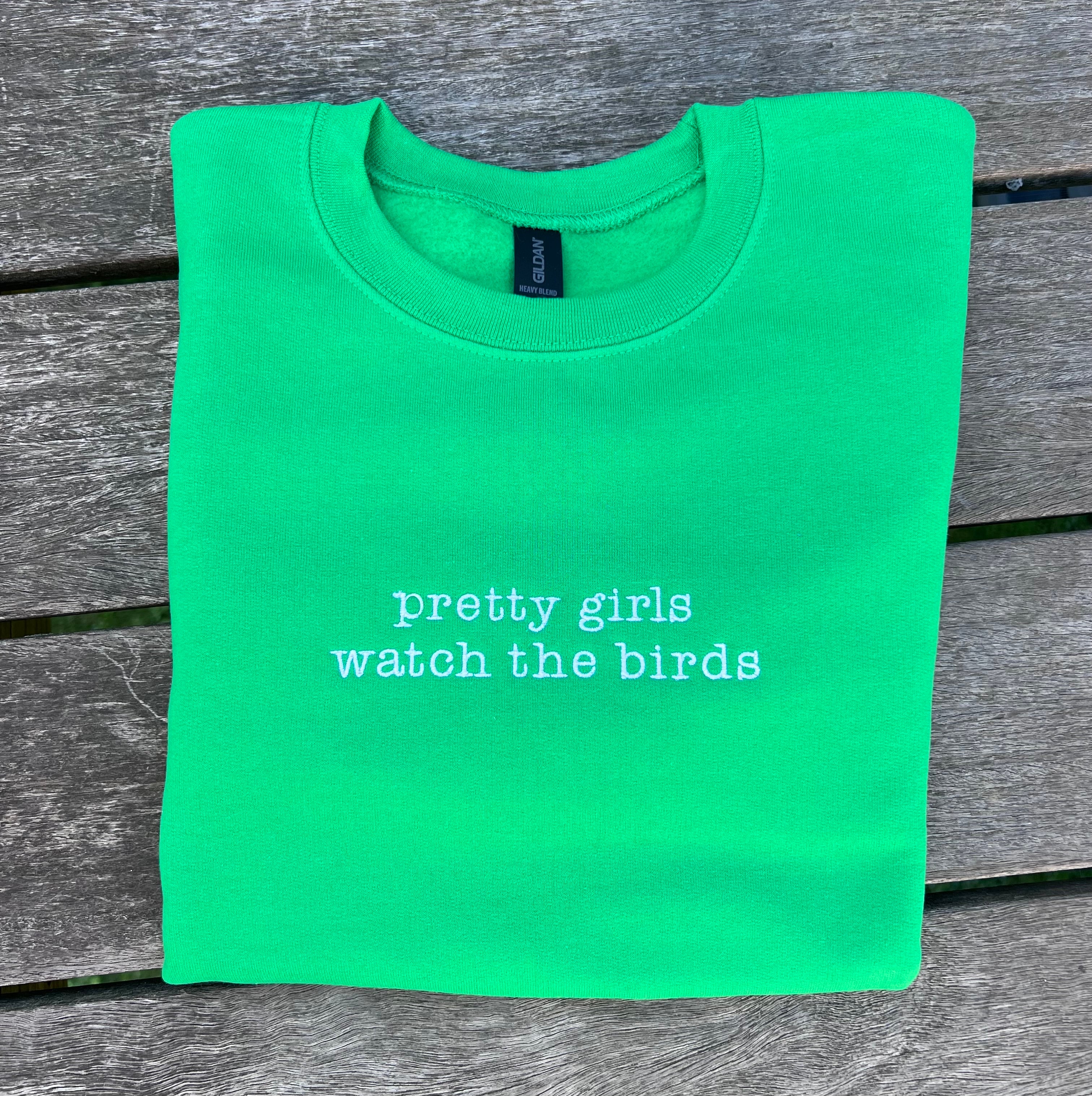 Green sweatshirt with 'pretty girls watch the birds' text on a wooden surface