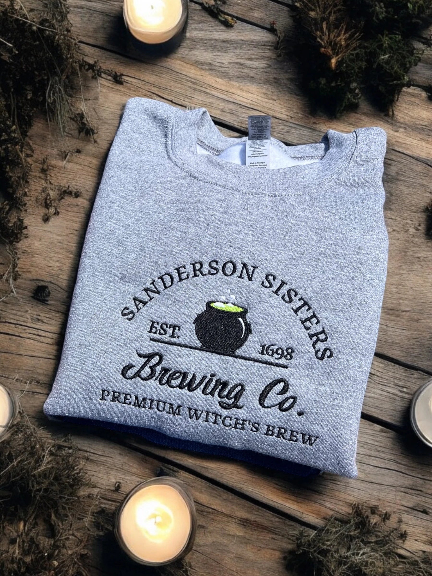 Gray sweatshirt with 'Sanderson Sisters Brewing Co.' text on a wooden surface with candles and herbs.